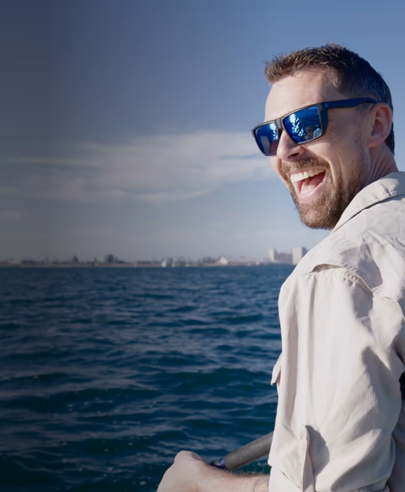 Smiling man wearing sunglasses and a light shirt, enjoying a sunny day on the water with a city skyline in the distance.