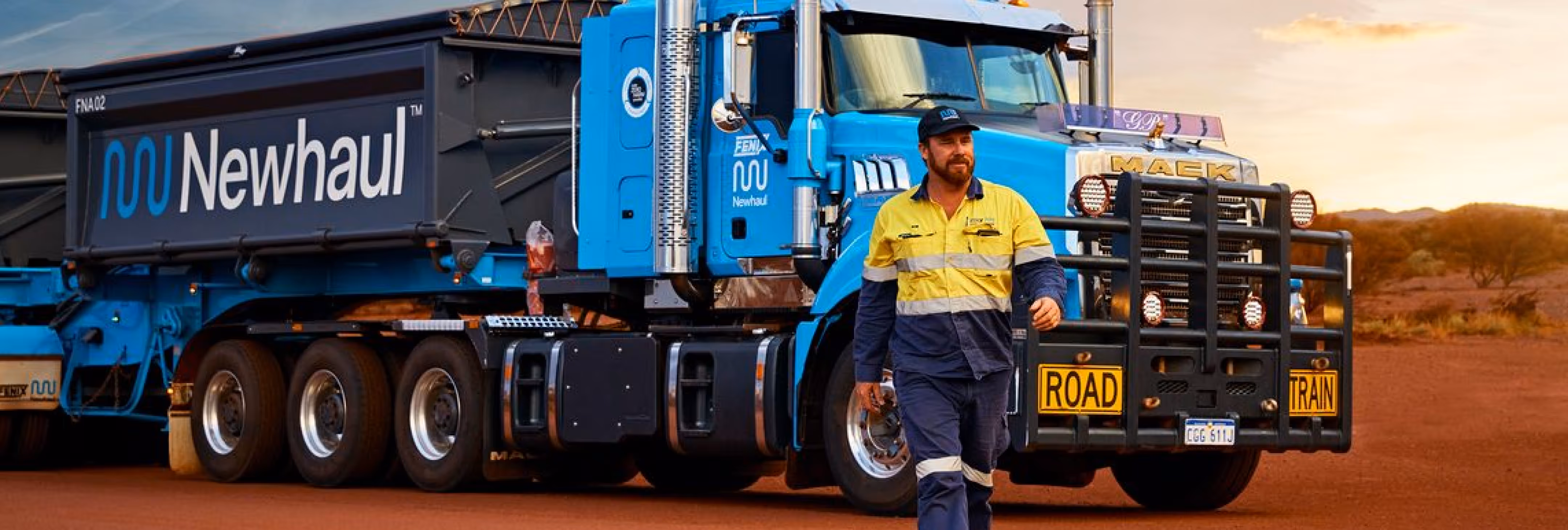 Man in high-visibility workwear walking in front of a blue Newhaul road train truck on a dirt road at sunset.