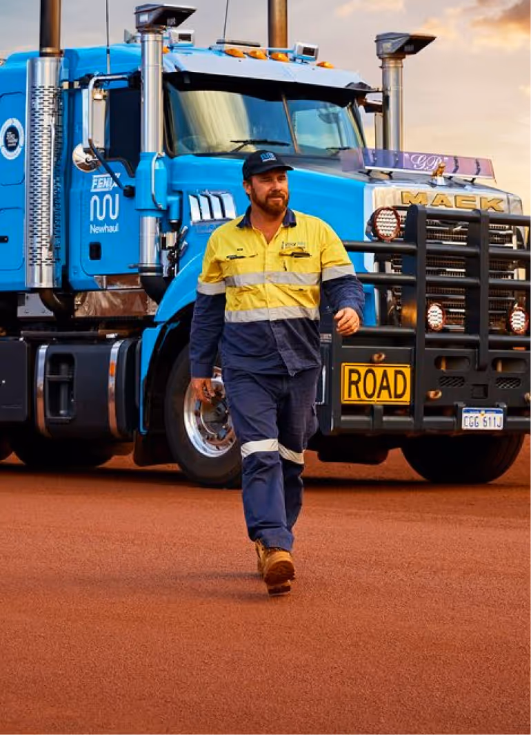 Man in high-visibility yellow and navy workwear walking on red dirt in front of a blue Mack truck with a bull bar and 'ROAD' sign.