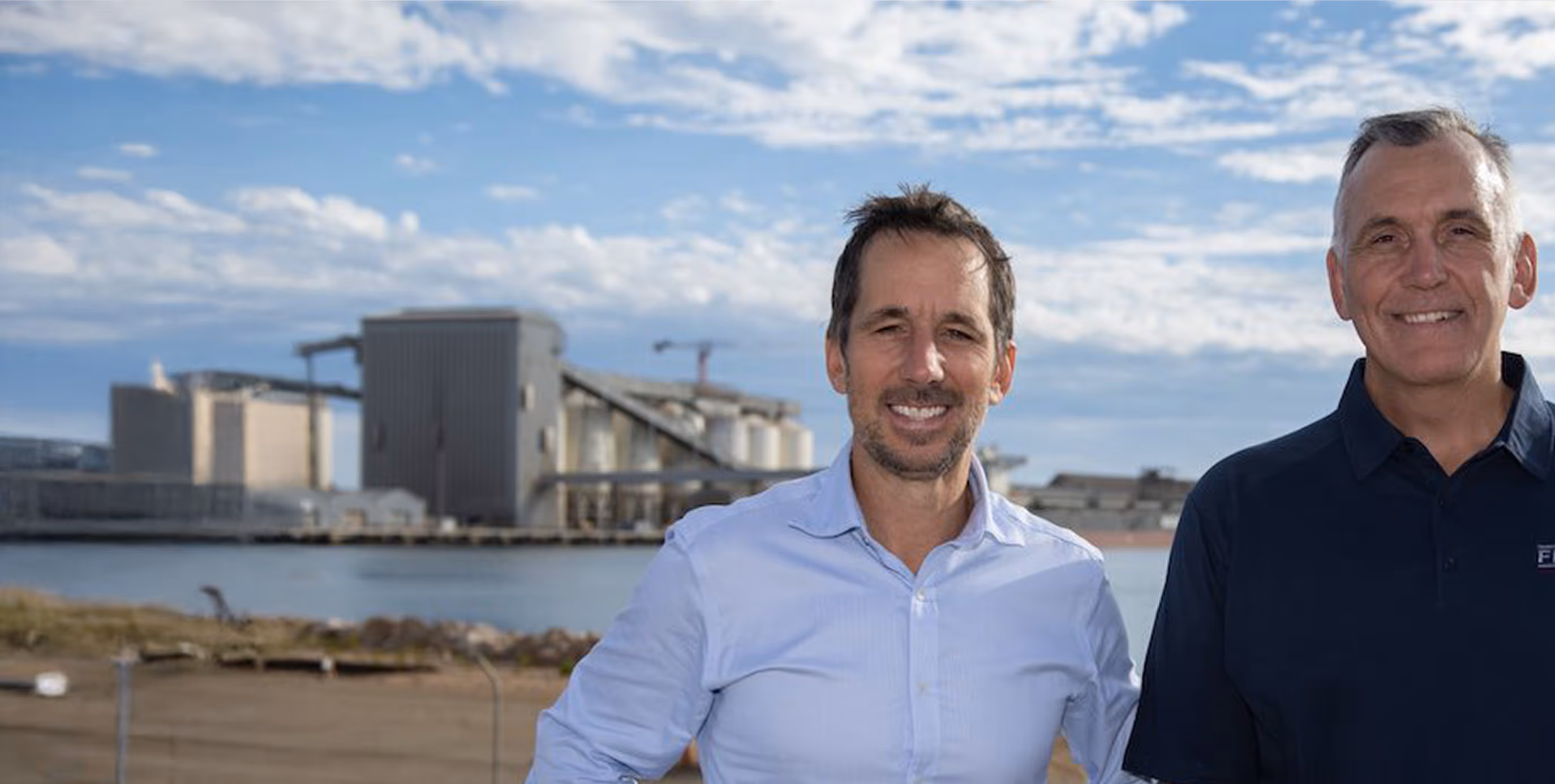Two men smiling and standing outdoors with an industrial plant and water in the background.