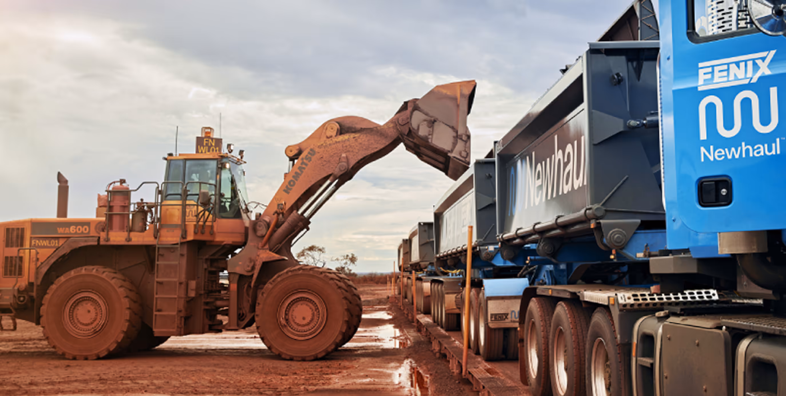 Rust-colored Komatsu WA600 loader filling blue Newhaul Fenix dump trucks on muddy construction site under cloudy sky.