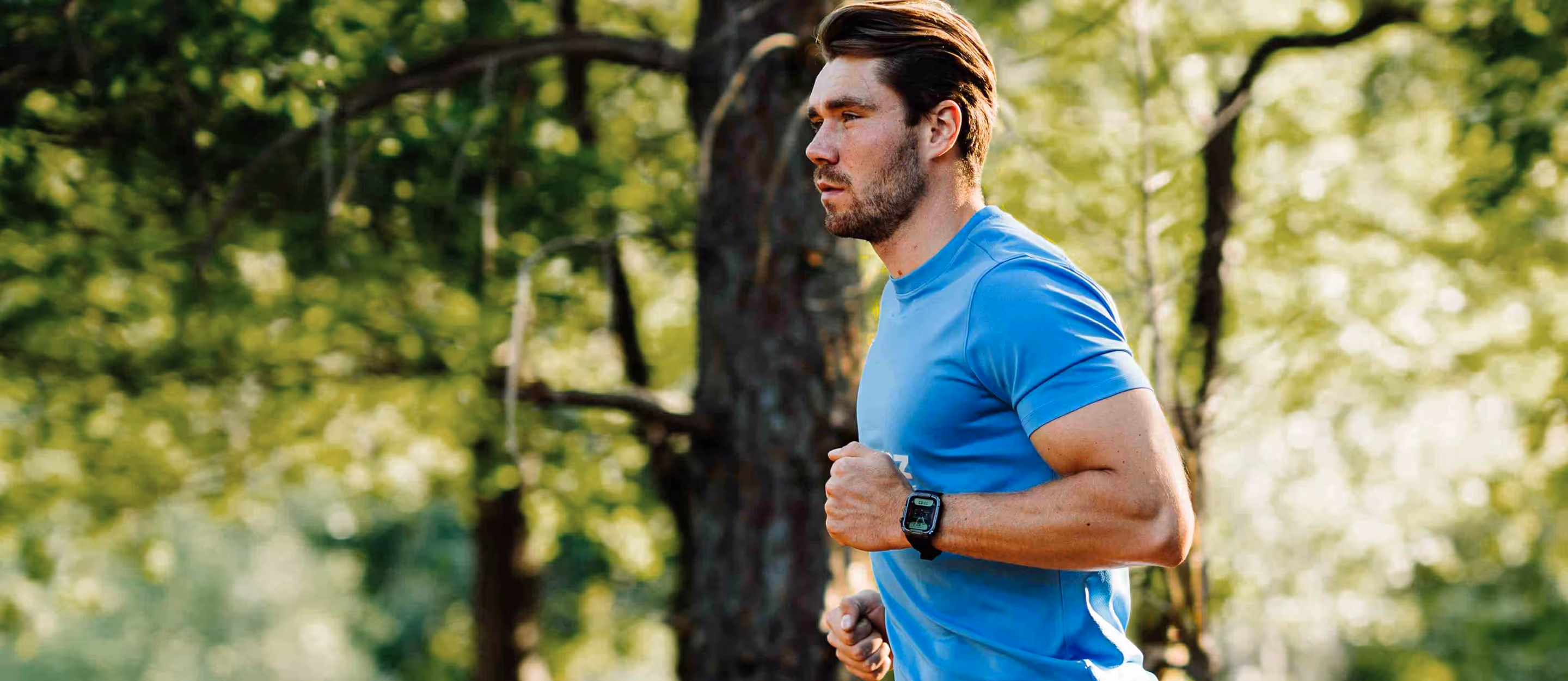Man jogging outdoors in a blue shirt with a smartwatch on his wrist, surrounded by green trees.