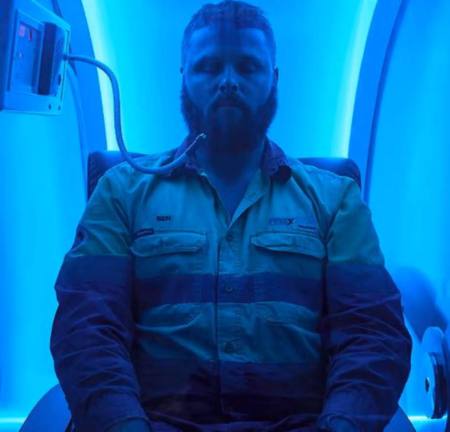 Bearded man in work uniform sitting inside a blue-lit chamber with an oxygen mask tube near his face.