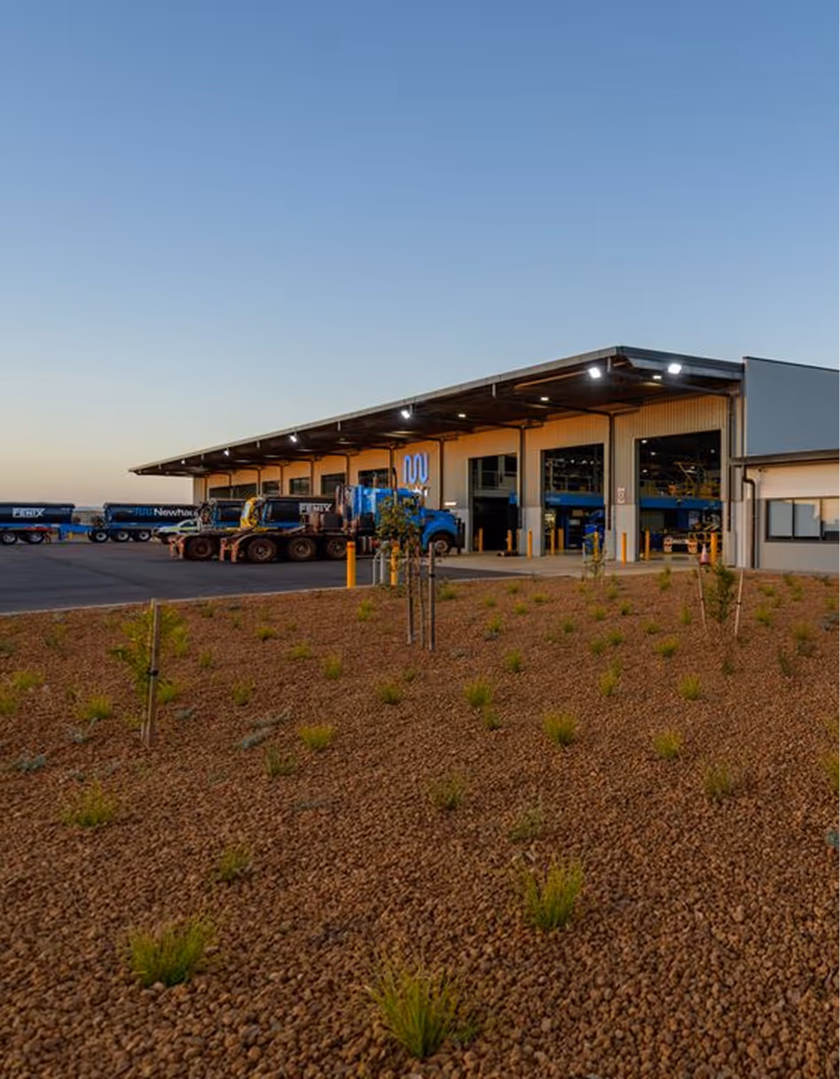 Modern industrial warehouse with several blue trucks parked outside under a clear sky at sunset.