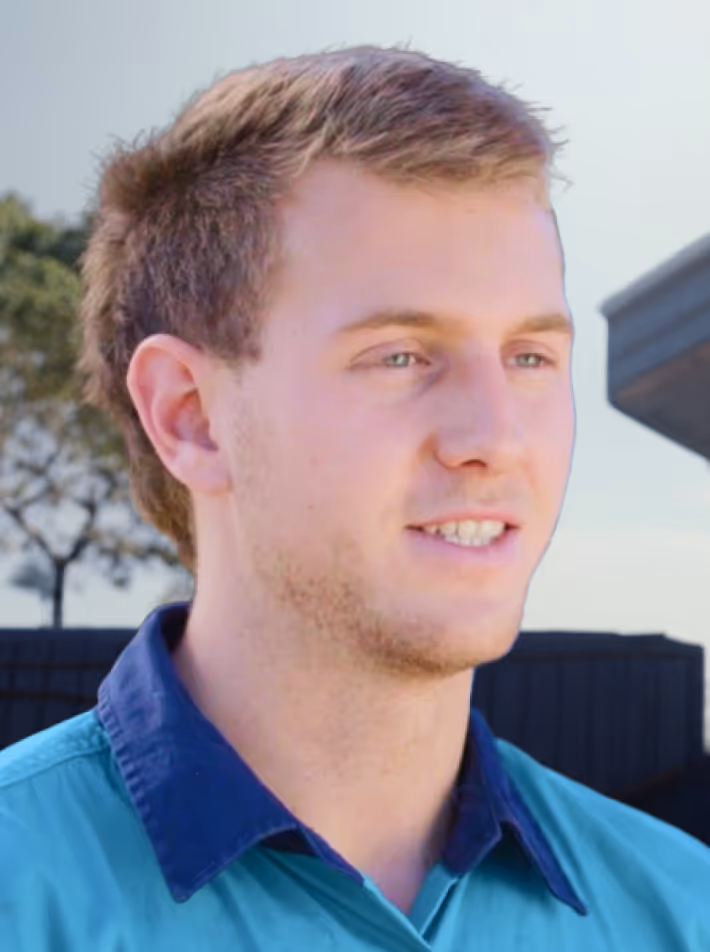 Young man with short brown hair wearing a teal shirt with a dark blue collar, standing outdoors with trees and a building in the background.