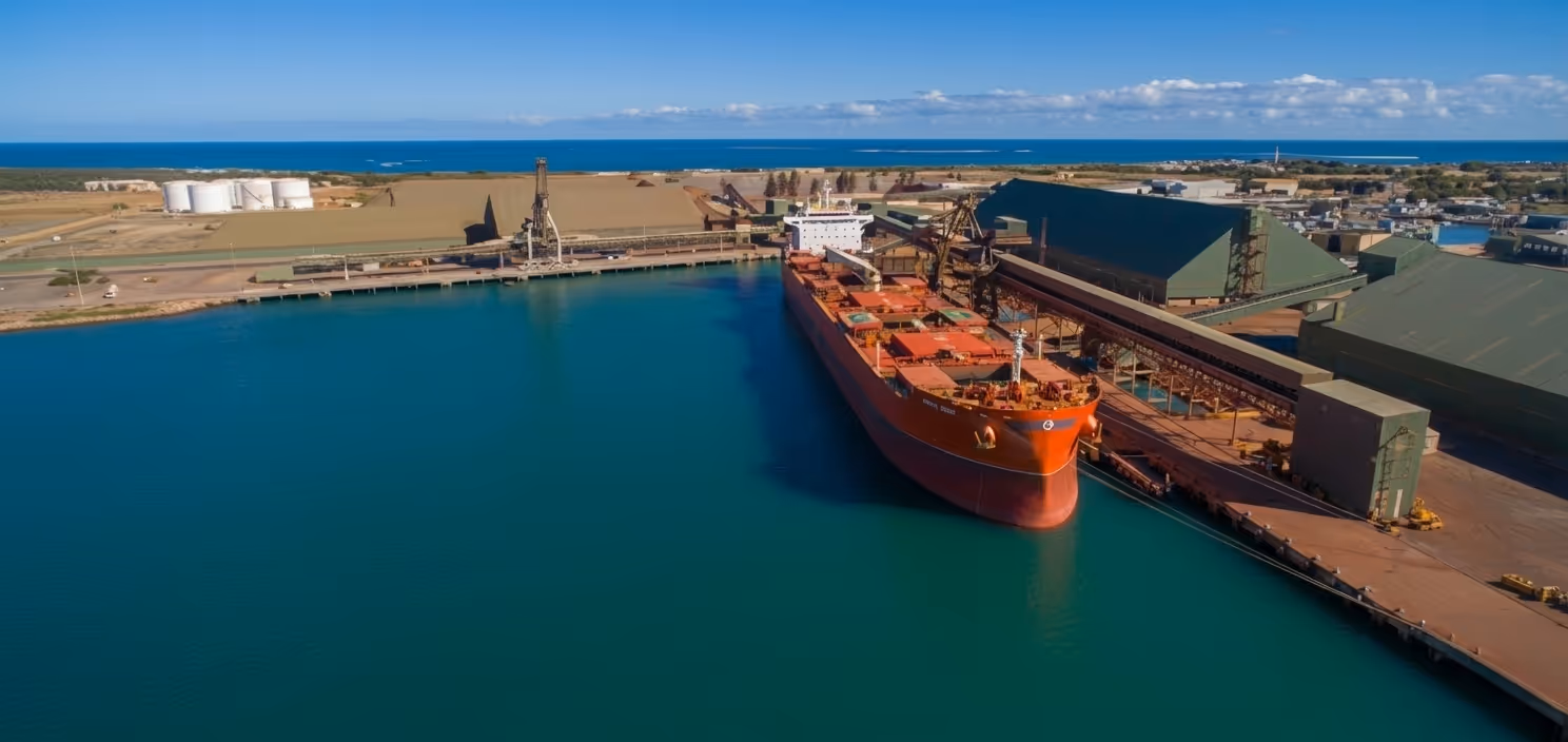 Large red cargo ship docked at an industrial port with warehouses and storage tanks near the ocean under a clear blue sky.