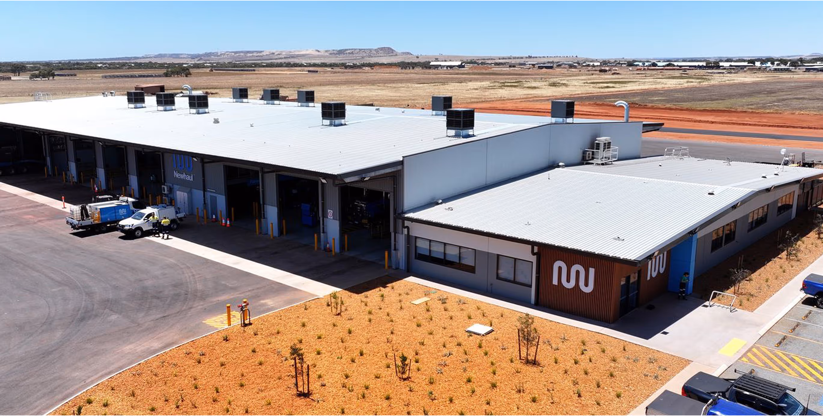 Aerial view of a large industrial workshop building with several garage bays, parked vehicles, and arid landscape in the background.