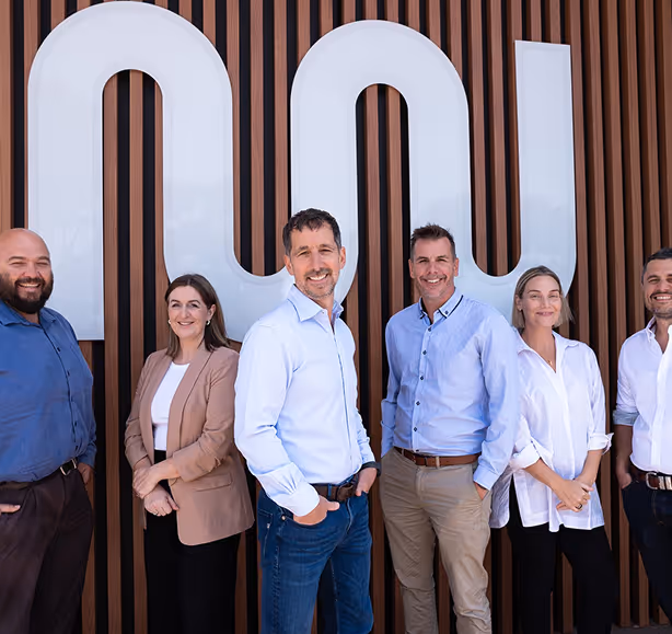 Six professionals standing and smiling in front of a large white Newhaul company logo on a wooden slat wall.