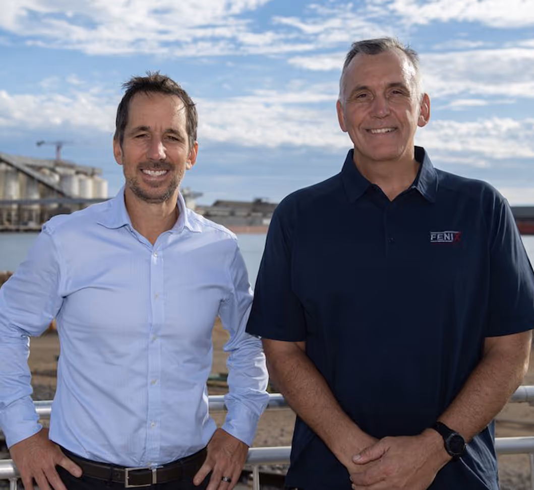 Two men (Craig Mitchell and John Welborn) standing outdoors near Geraldton Port with industrial buildings and a partly cloudy sky in the background, both smiling at the camera.