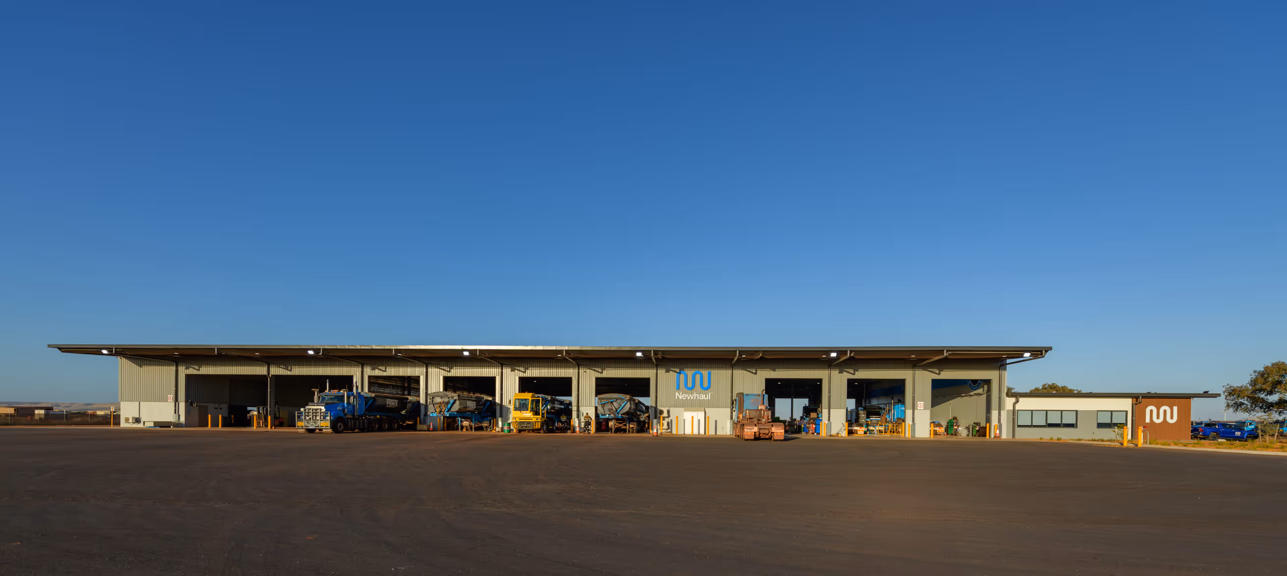 Wide view of Newhaul's industrial workshop building with several trucks and heavy equipment parked inside and outside under a clear blue sky.