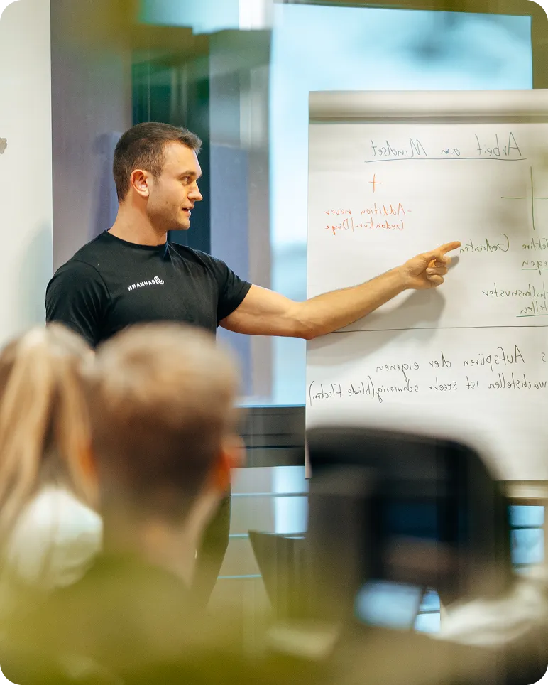 Mann in schwarzem T-Shirt, der während einer Präsentation vor Publikum auf eine weiße Tafel zeigt.