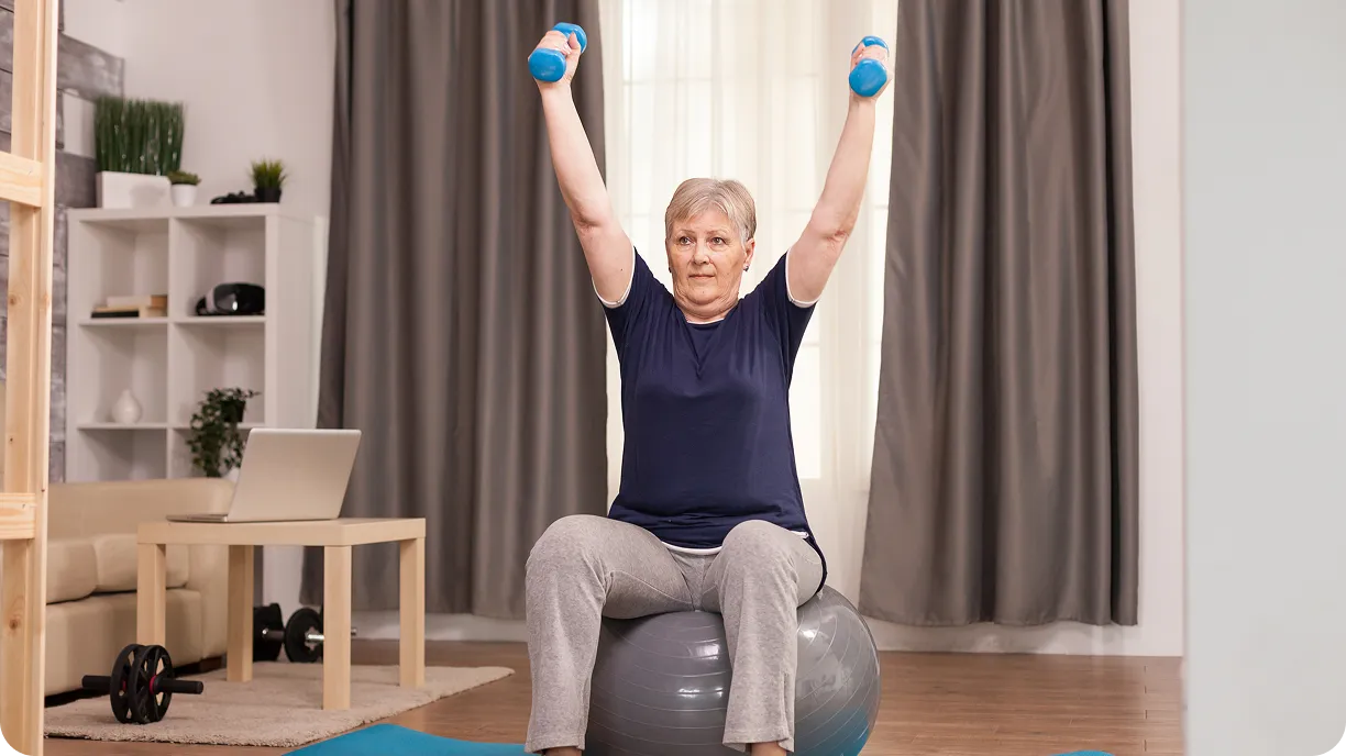 Ältere Frau, die mit blauen Hanteln trainiert, während sie auf einem grauen Gymnastikball im Wohnzimmer sitzt.