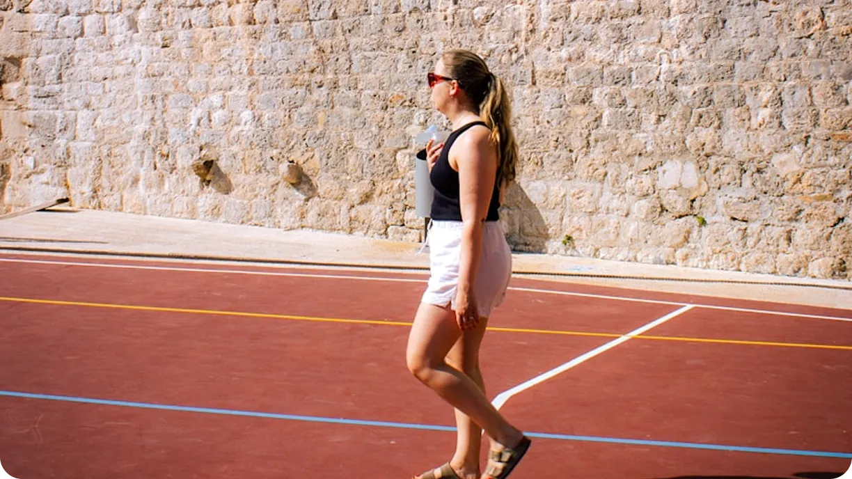 Frau in schwarzem Tanktop, weißen Shorts und Sandalen, die auf einem Outdoor-Sportplatz mit einer Steinmauer im Hintergrund spazieren geht und eine Wasserflasche in der Hand hält.