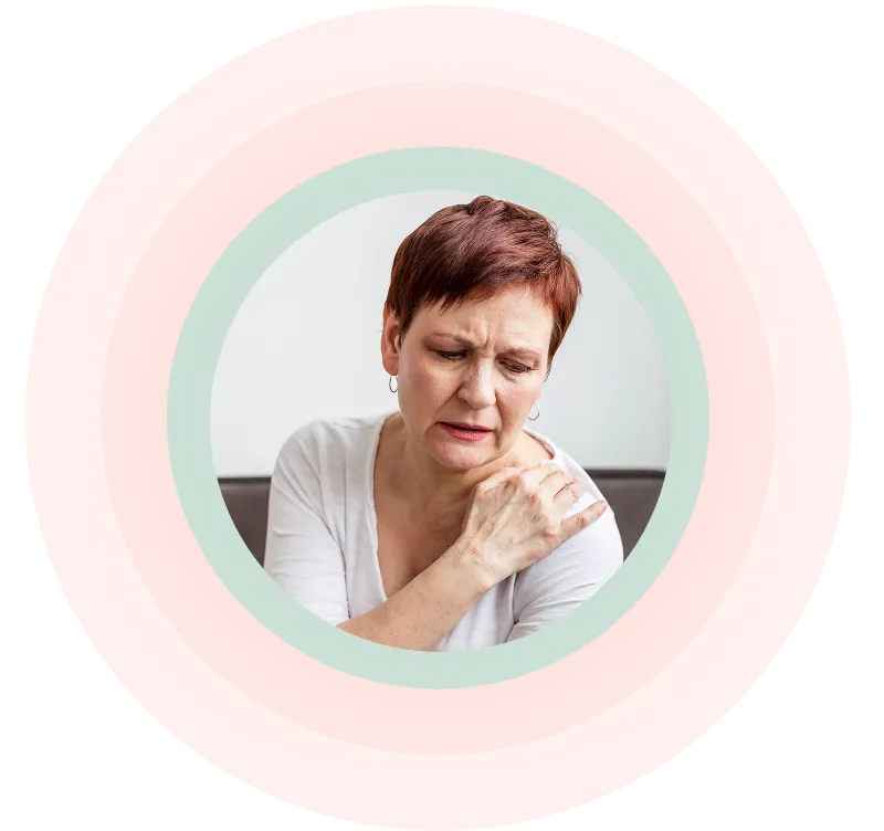 Eine Frau mittleren Alters mit kurzen Haaren, die ein weißes Hemd trägt und sich mit schmerzverzerrtem Gesicht die Schulter hält.