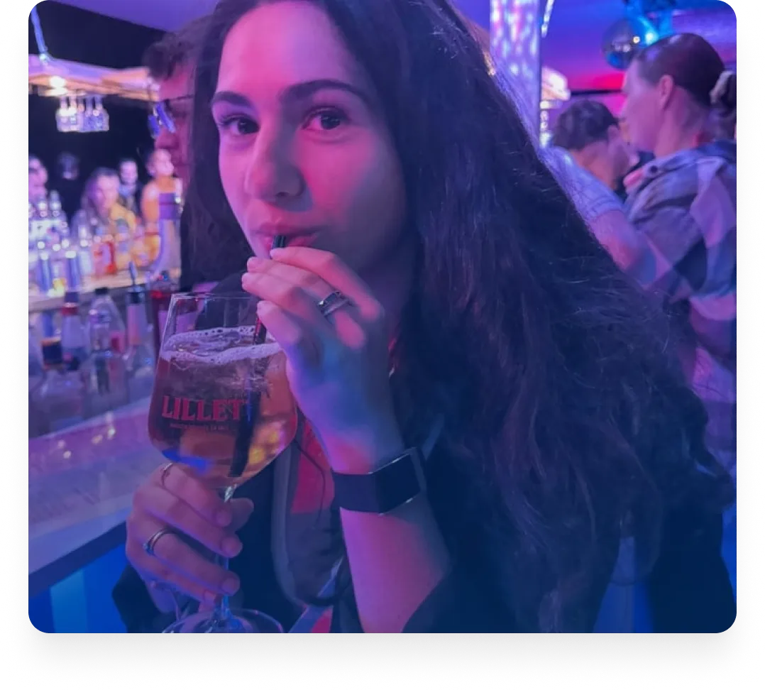 Frau mit langen dunklen Haaren, die in einer Bar mit bunter Beleuchtung mit einem Strohhalm an einem Getränk nippt.