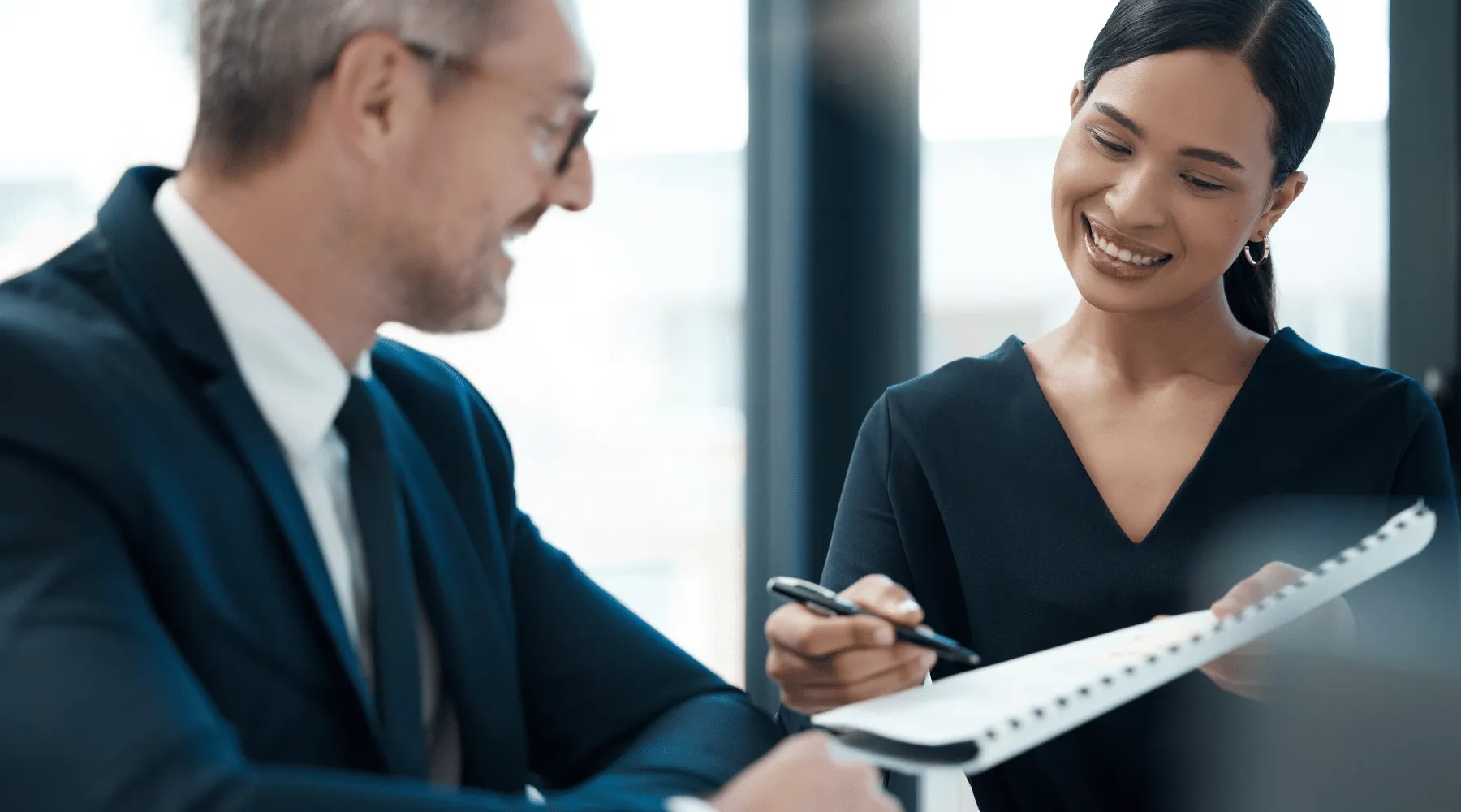 A man in a suit and a woman in a business dress stand side by side, engrossed in reviewing a notebook that the woman holds. As she points at a detail in the notebook with her pen, their focused expressions and collaborative posture exemplify the principles of corporate training on accountability. This scene illustrates the importance of shared responsibility, clear communication, and active engagement in tasks, highlighting how accountability forms a cornerstone of effective teamwork and professional integrity in the workplace.