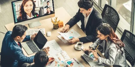 A scene of four people seated around a table, deeply engaged in a meeting that also includes a group of remote participants displayed on a monitor. This setup exemplifies corporate training on effective meetings, highlighting how to seamlessly integrate in-person and virtual attendees. The image underscores the importance of inclusive, well-structured, and productive meetings in a modern, hybrid work environment, demonstrating the effectiveness of combining face-to-face and remote communication for collaborative success.