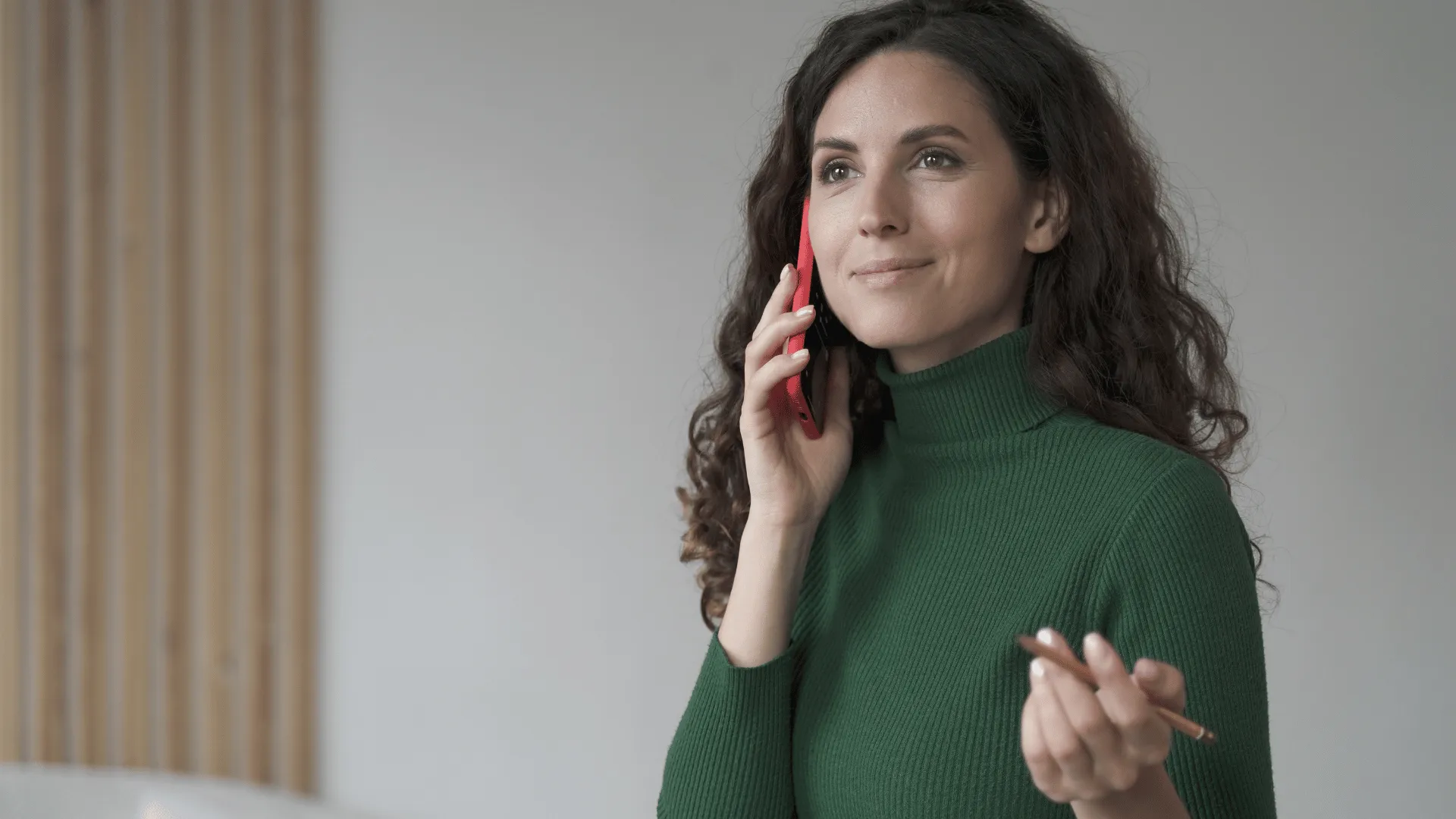 A woman in a green turtleneck sweater with long, curly brown hair is standing while talking on a phone. She's holding it up to her right ear. In her left hand, she holds a pencil. She was just reviewing the learning KPIs provided by Electives, and she’s feeling good about her decision to outsource L&D.