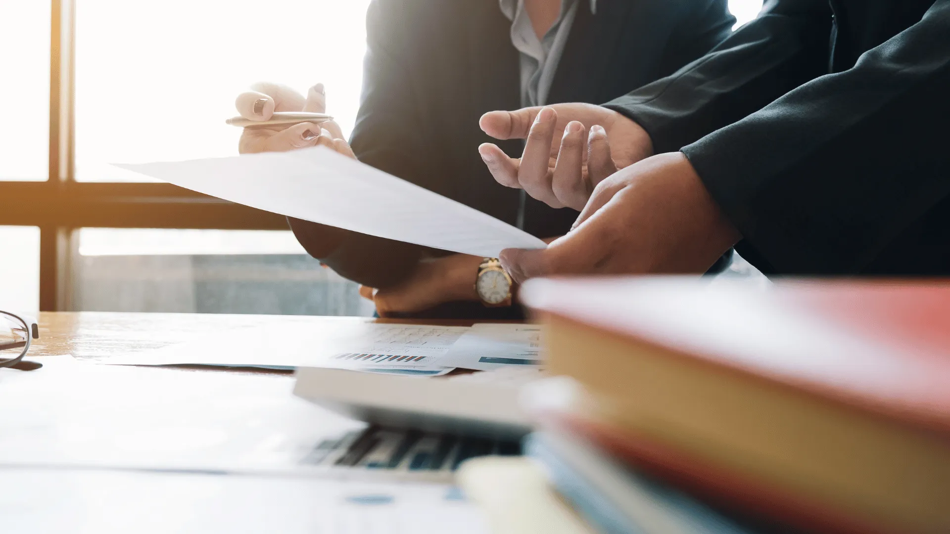 A close up picture of 2 people (a man and a woman) engaged in a conversation at work. We see only their hands and parts of their upper body. The man is holding a piece of paper and the woman is holding a pen.