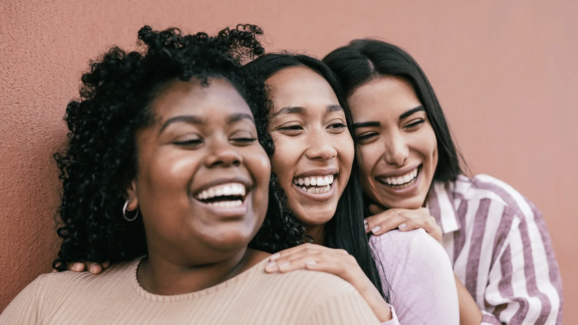 3 women of color standing near each other and smiling. We see them only from their shoulders up. A pinkish/tanish concrete wall is behind them.