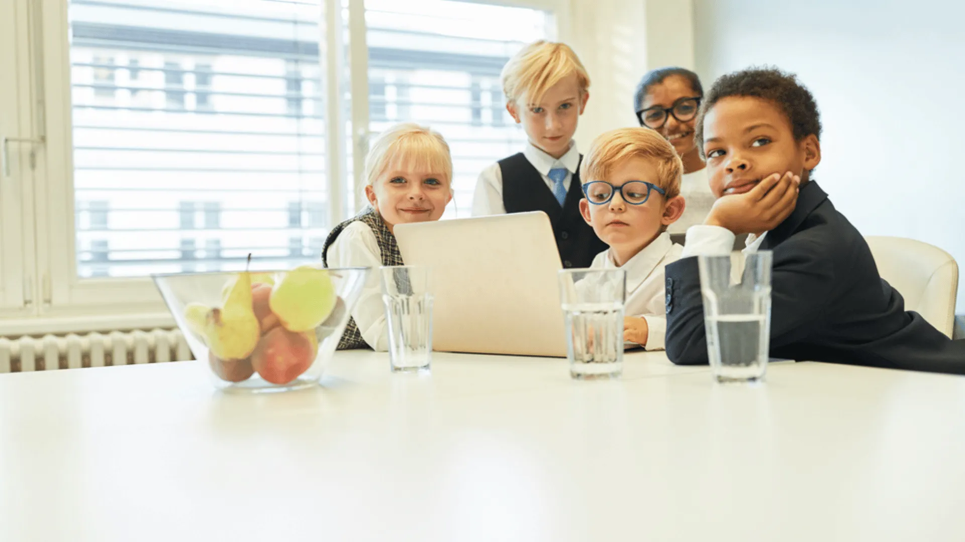 A group of 5 children dressed in business attire are gathered at the end of a conference room table. There is an open computer in front of one kid and the rest are around him. Their facial expressions range from happy to amused.