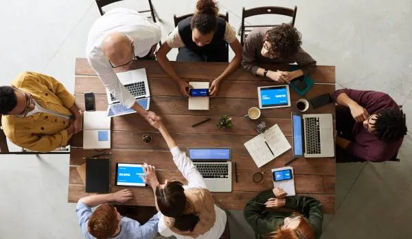 Overhead shot looking down at a table of diverse people working on their laptops, smiling and shaking hands while reviewing data.