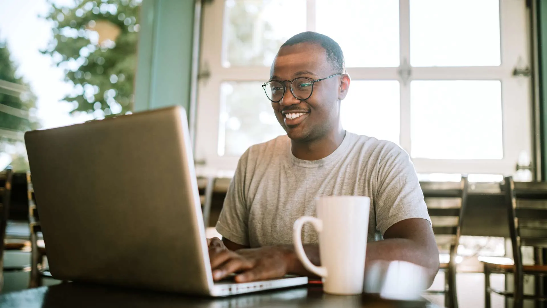 A young man with glasses works on his laptop from a coffee shop, smiling.