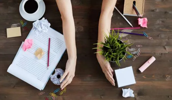A messy desk being cleaned off. A woman’s arms are in the middle of the desk, facing outward, as if they’re about to sweep everything off to the sides of the desk.