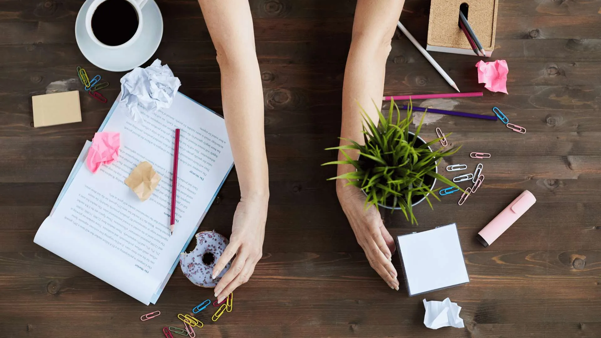 A messy desk being cleaned off. A woman’s arms are in the middle of the desk, facing outward, as if they’re about to sweep everything off to the sides of the desk.