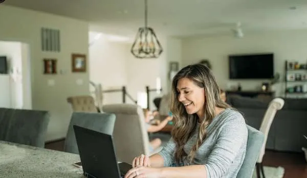 A woman in her early 40s works from her kitchen island. Behind her, her home is blurred.