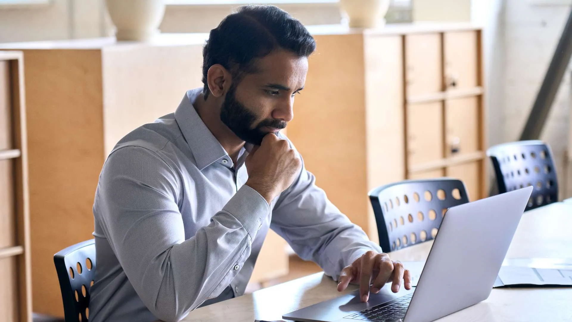 A man with a beard rests his chin on his hand as he looks at something intently on his laptop.