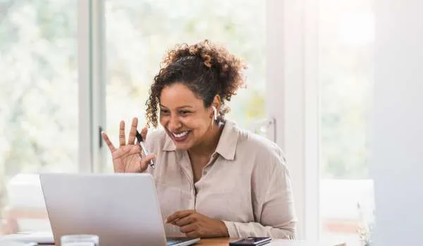 A middle-aged woman is smiling and waving at her laptop as she joins a live virtual training.