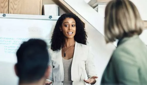 A young black woman is presenting to two colleagues. We are looking through the shoulders of the colleagues at her and her whiteboard.