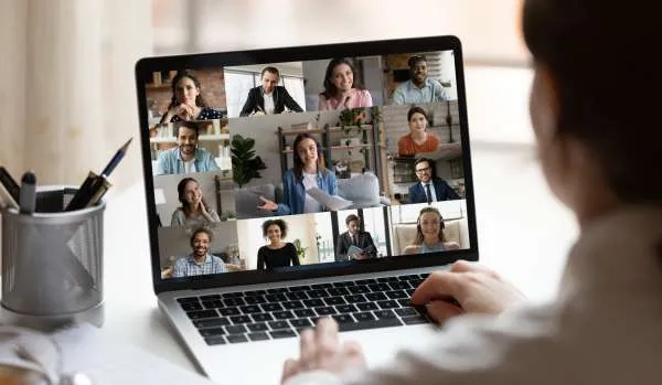 We are looking over a man's shoulder at his laptop on his desk. He is on a virtual training with a diverse group of people.