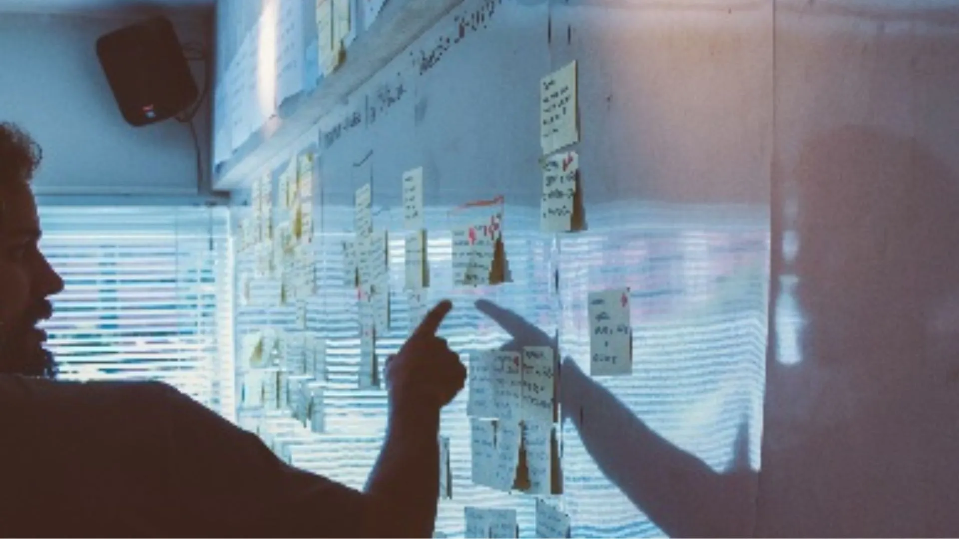 A person stands in front of a wall covered with sticky notes and charts during a corporate training session for strategic planning. The individual appears to be in deep thought or explanation, pointing at specific items that seem to outline a strategy or plan, with the shadow of their hand cast prominently on the board. The ambient lighting suggests a focus on the task at hand, underscoring the importance of careful planning in a business context.