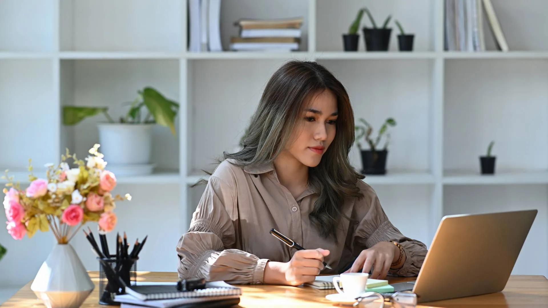A young woman is looking at her laptop and taking notes with a pen and notebook.