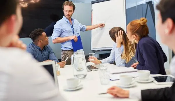A man is leading a workshop in a conference room. A group of people is looking at him as he points at the flipchart.
