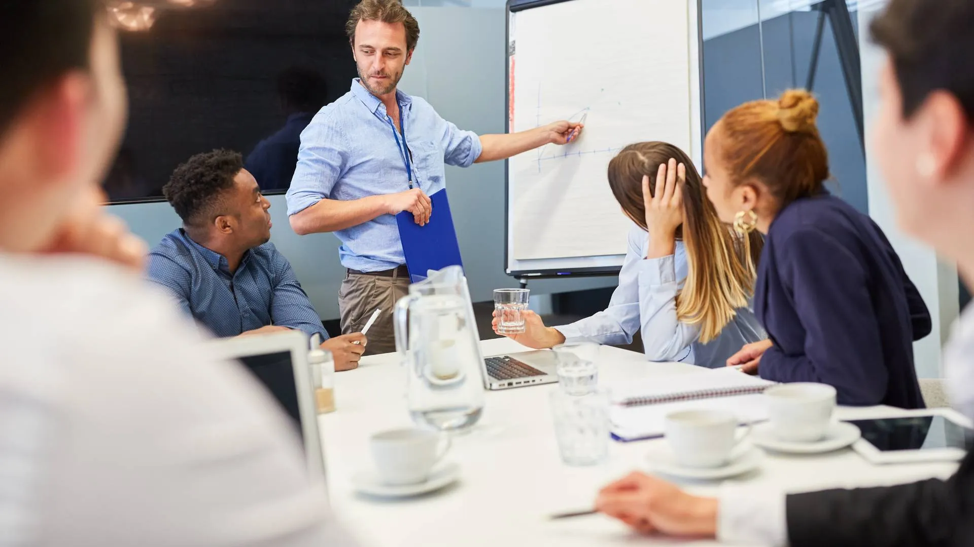 A man is leading a workshop in a conference room. A group of people is looking at him as he points at the flipchart.