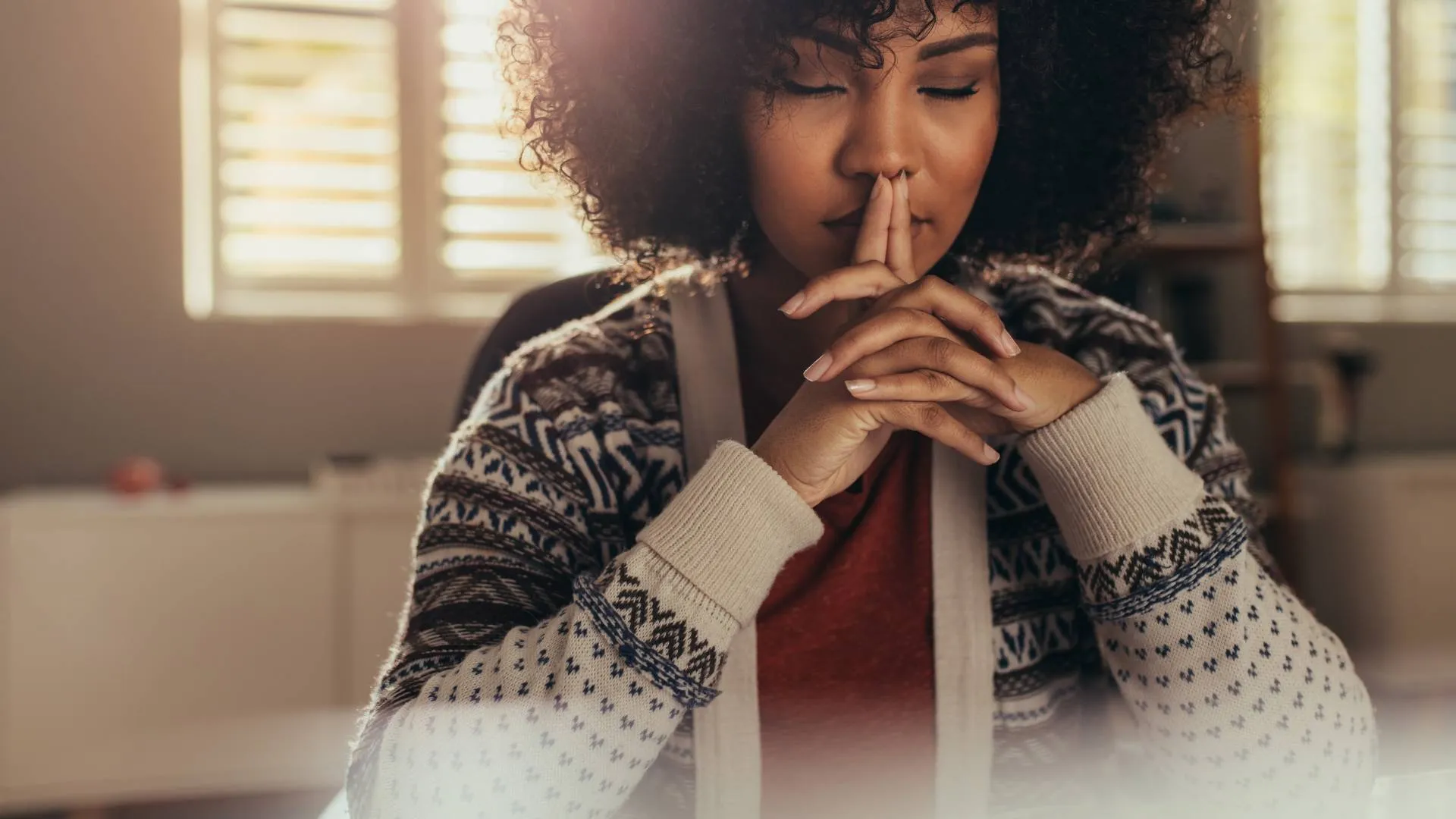 A woman sits with her elbows on the table, hands clasped together. Her eyes are closed as she rests her chin on her hands.