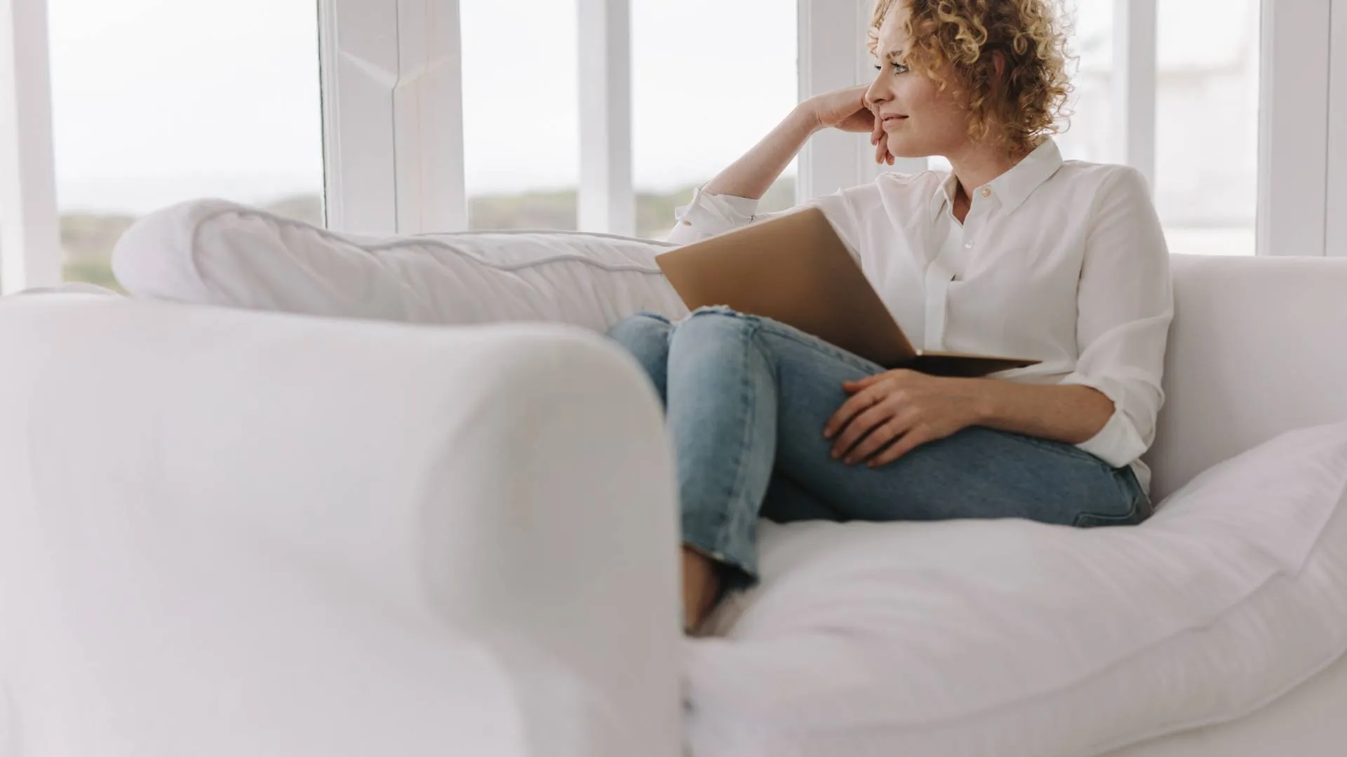 A woman is sitting on her couch, laptop in her lap, while looking out the window.