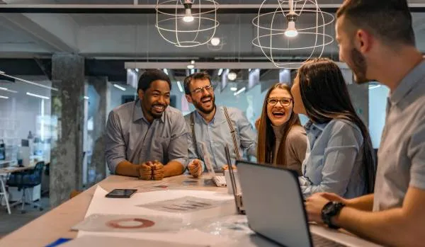 A diverse group of employees is standing at a common work area, laughing and smiling.