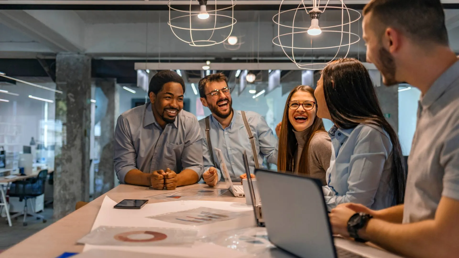 A diverse group of employees is standing at a common work area, laughing and smiling.