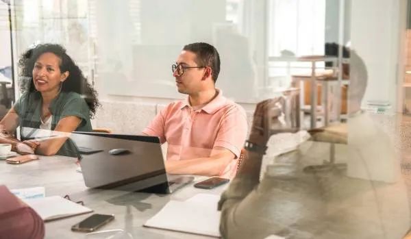 We are looking through a glass conference room wall at a diverse group of people participating in a training around a conference table.