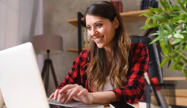 A young woman smiles while working from her home office on her laptop.