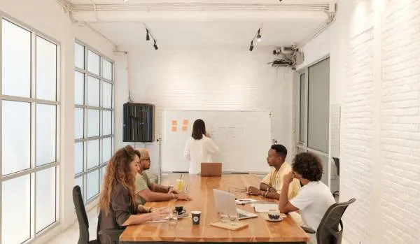 A diverse group of people are meeting in a modern conference room. Four are sitting at a conference table discussing while one is writing on the whiteboard.