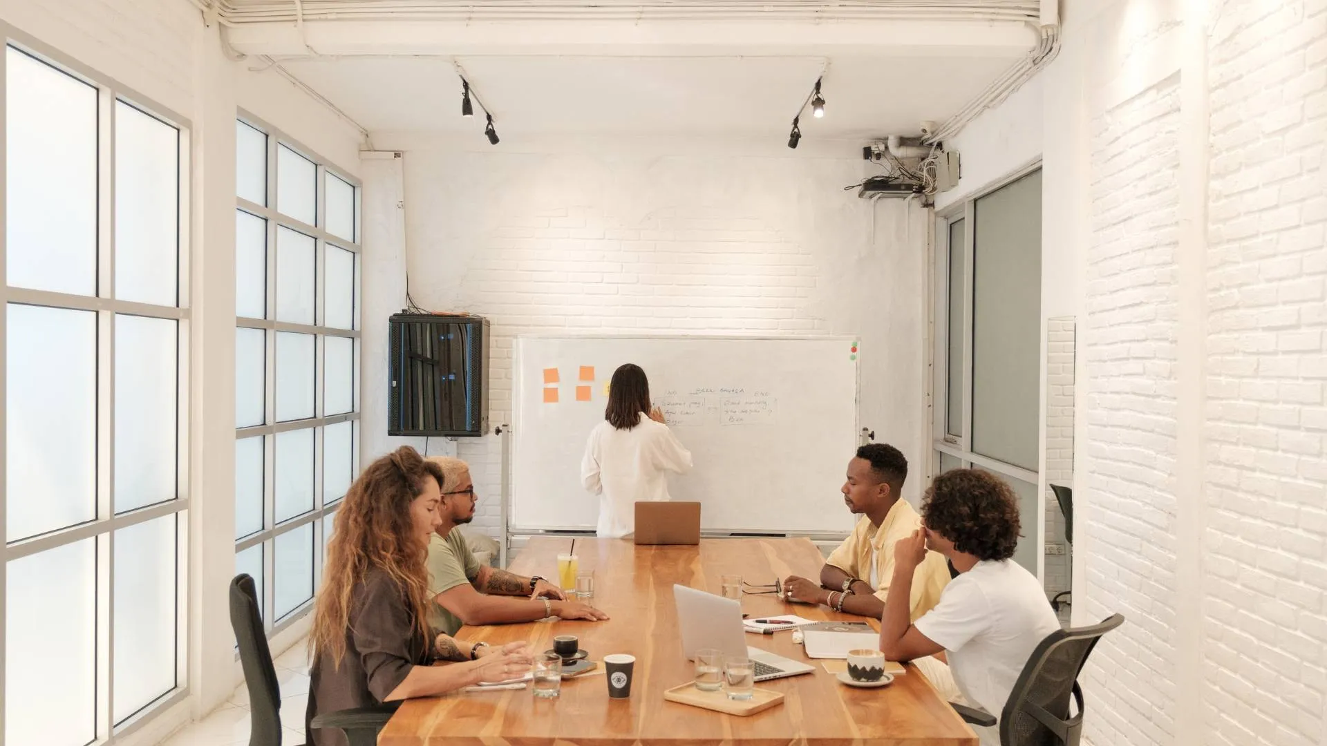 A diverse group of people are meeting in a modern conference room. Four are sitting at a conference table discussing while one is writing on the whiteboard.