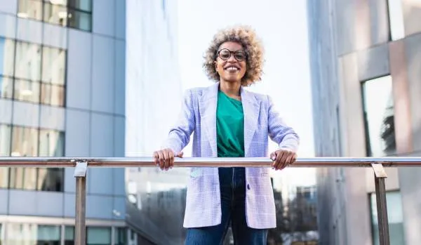 A smiling woman in glasses is looking at us from an elevated walkway, holding onto the railing.