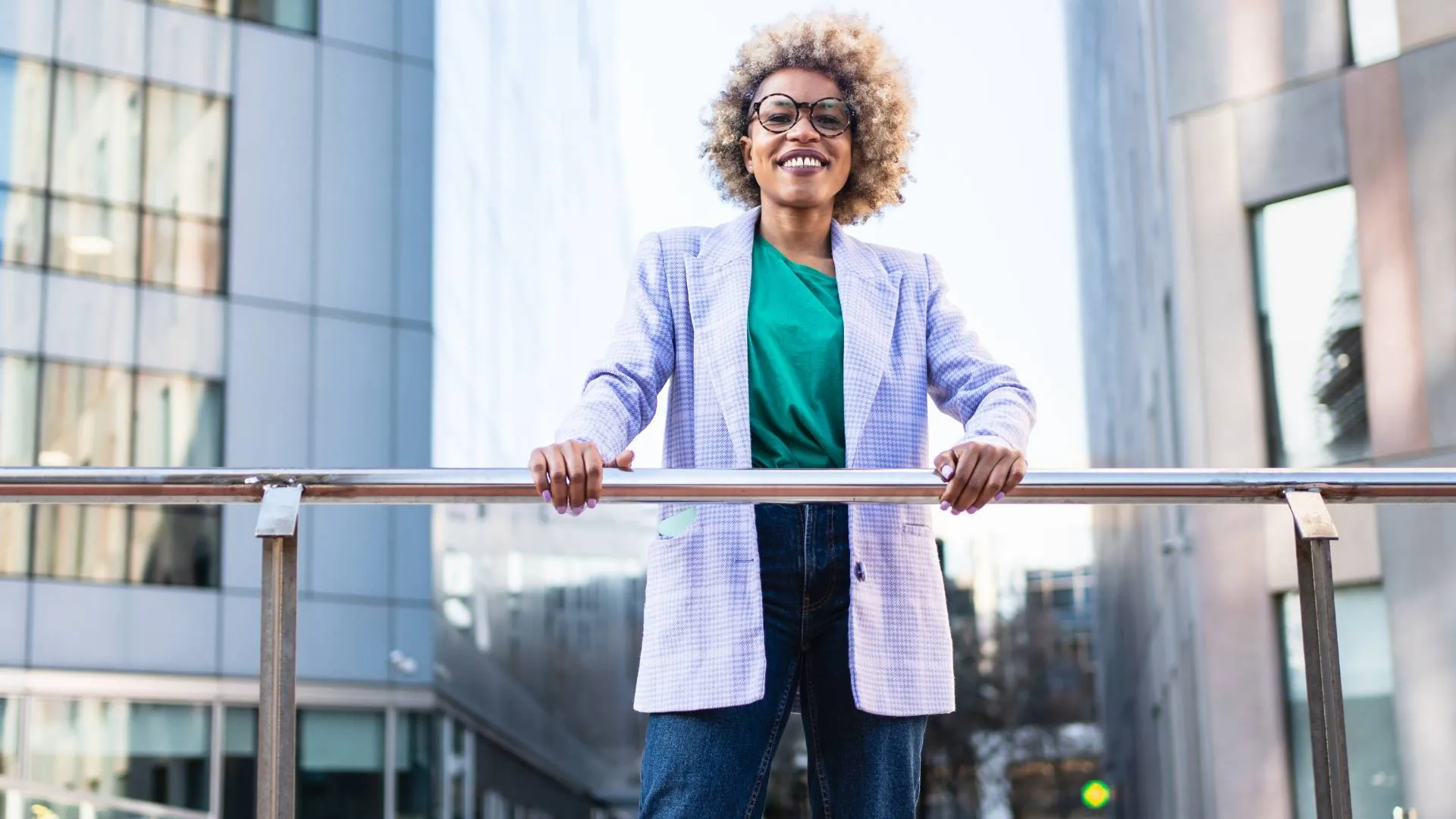 A smiling woman in glasses is looking at us from an elevated walkway, holding onto the railing.