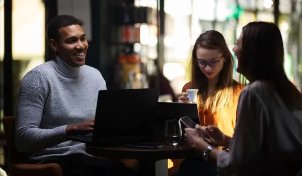 A group of three young professionals are working together in a coffee shop and laughing.