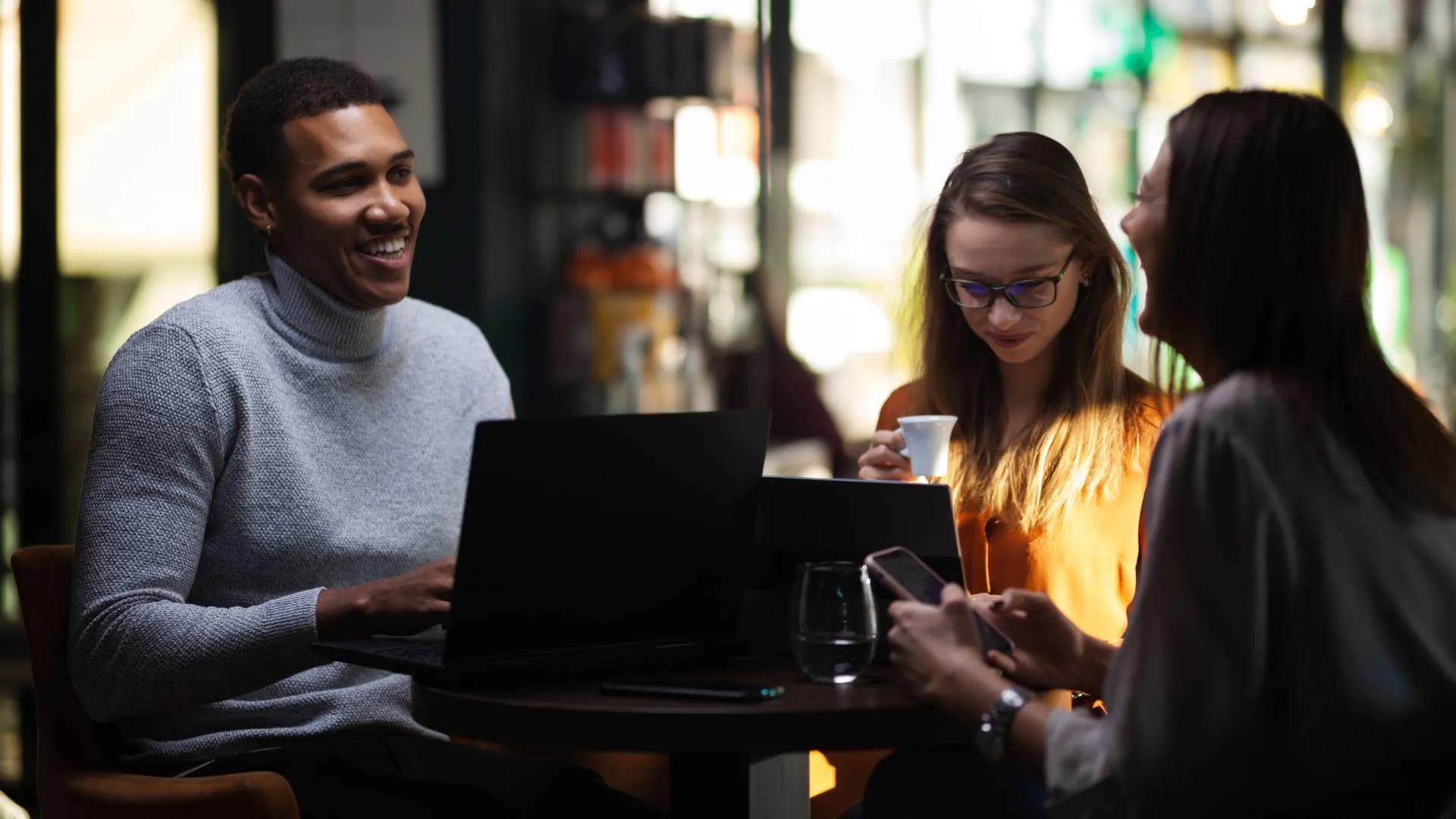 A group of three young professionals are working together in a coffee shop and laughing.